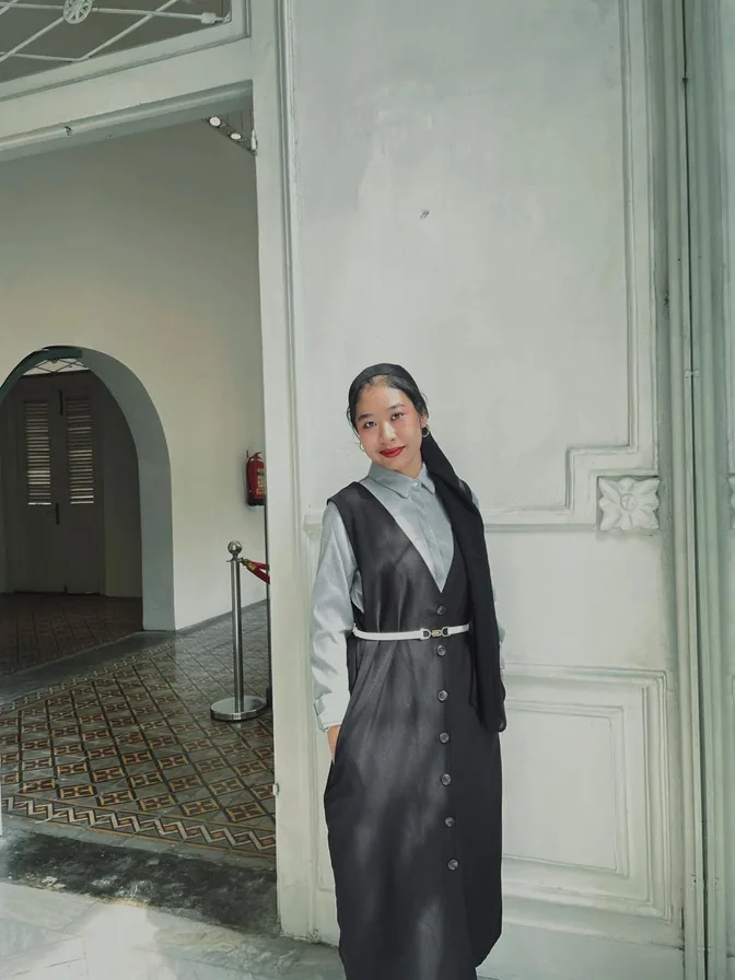 Alika standing by an arched doorway in a dark pinafore dress